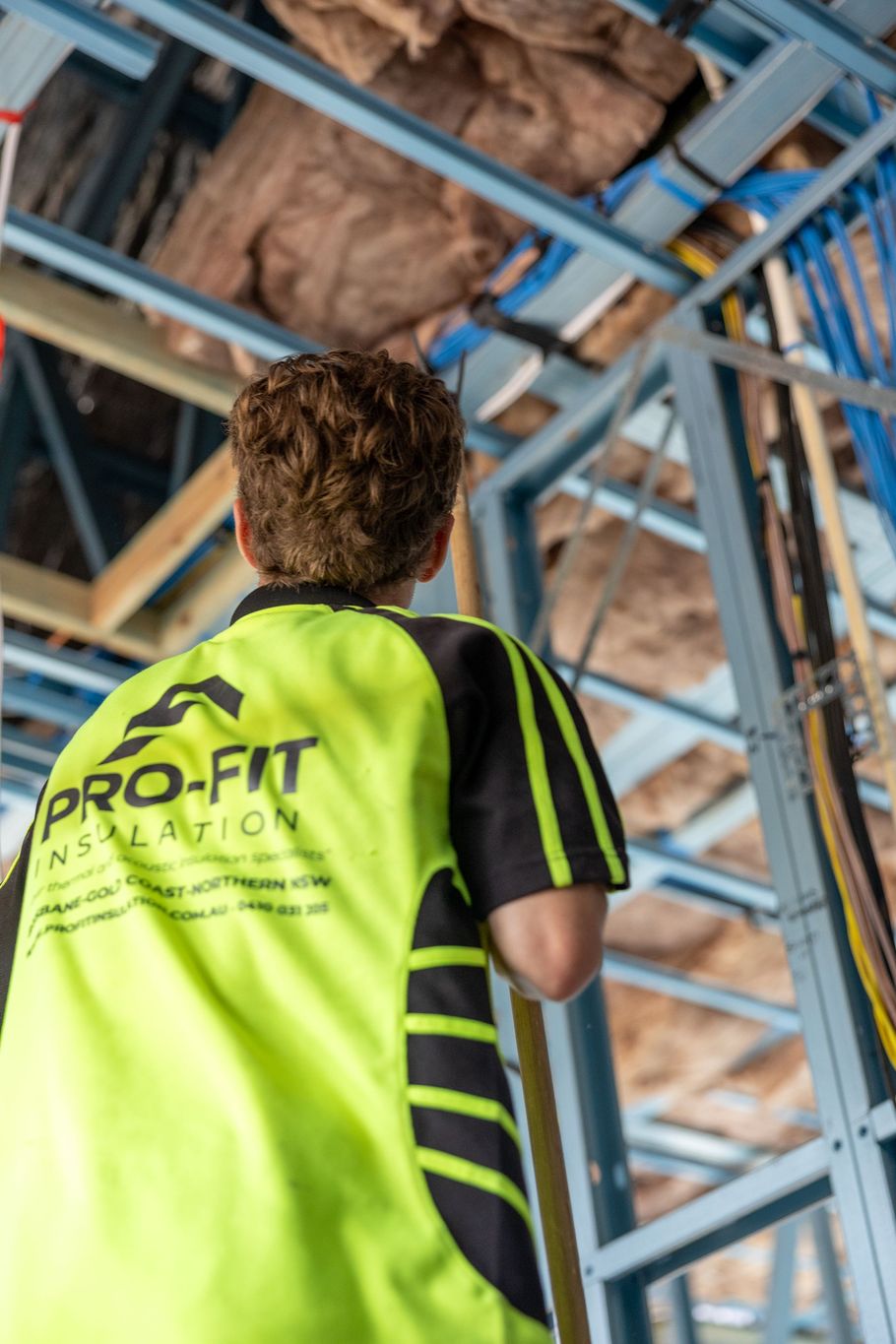 Construction worker in a lime green shirt with tool belt, drilling into wooden ceiling beams.