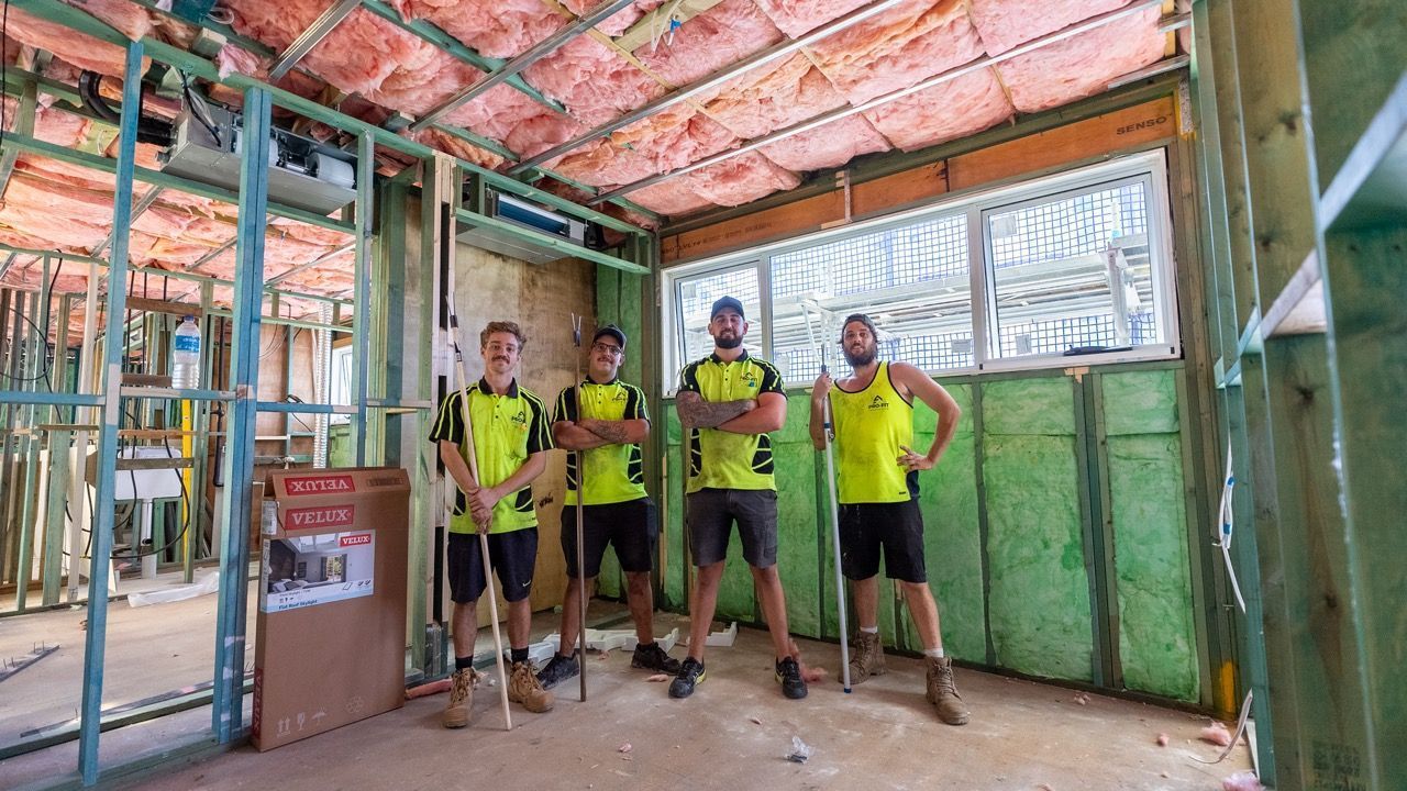 Four workers in construction zone; yellow shirts, arms crossed, unfinished walls, pink insulation.