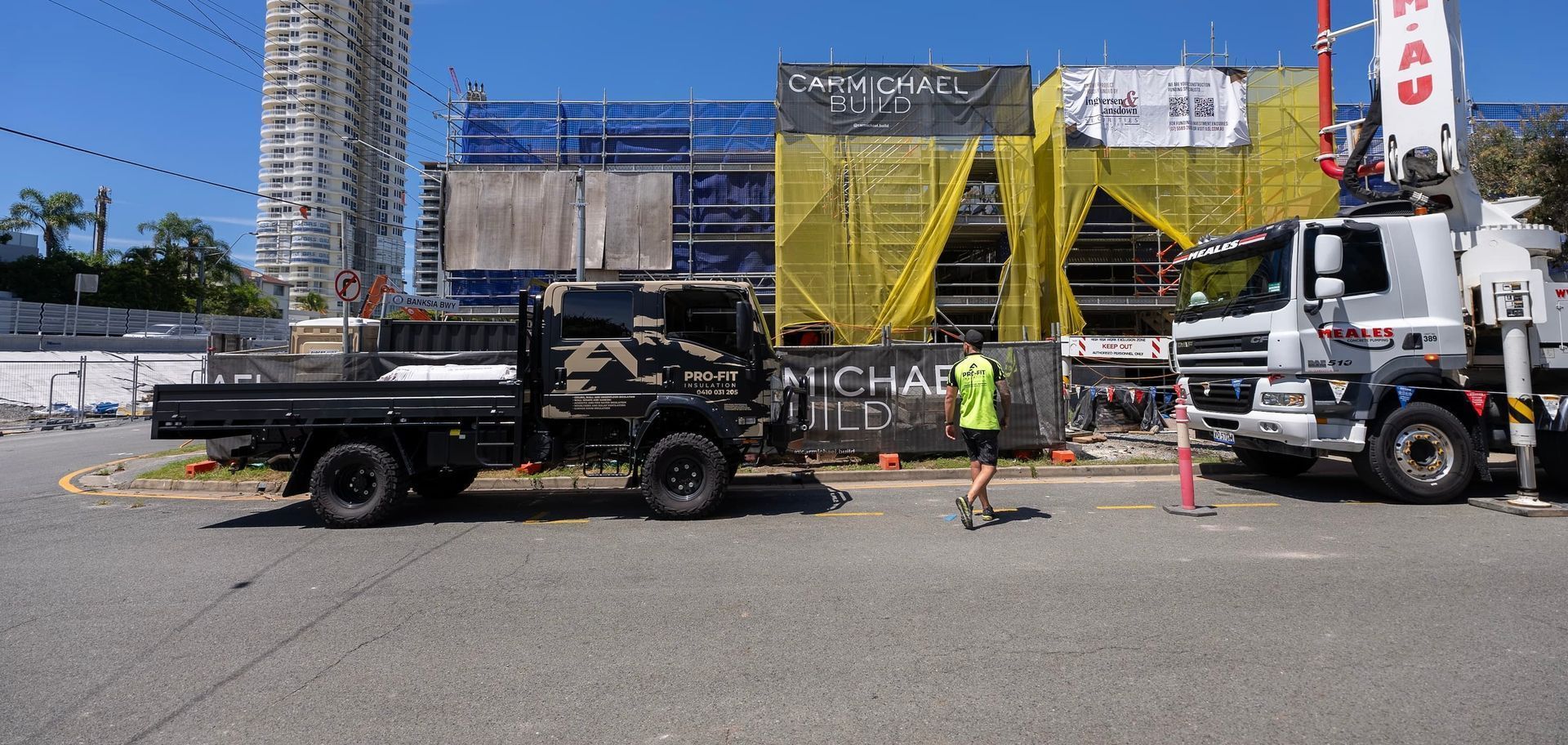 Construction site with trucks, a person in a vest, and buildings against a blue sky.