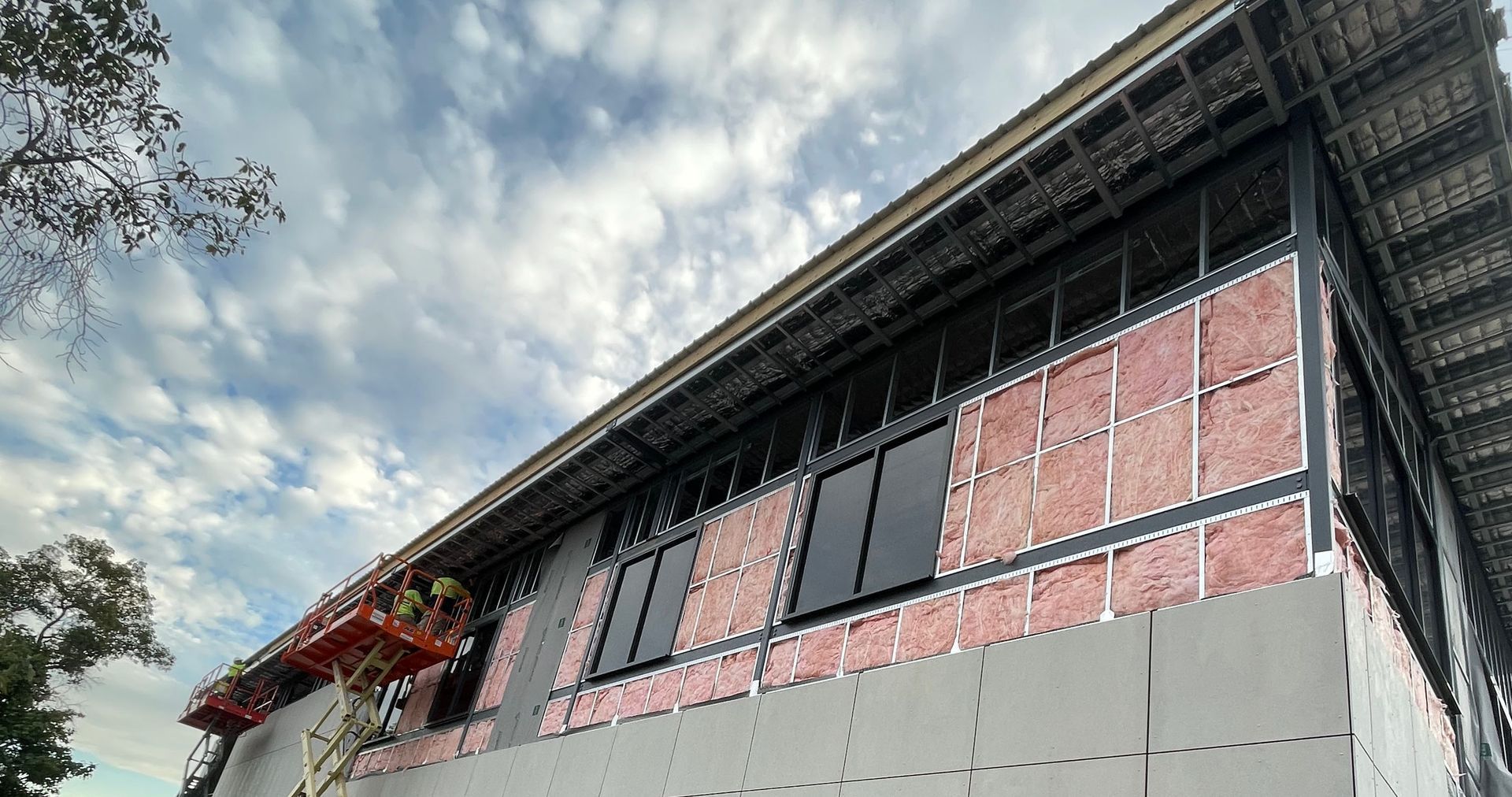 Building exterior under construction; pink insulation, black window frames, gray panels, workers on lift.