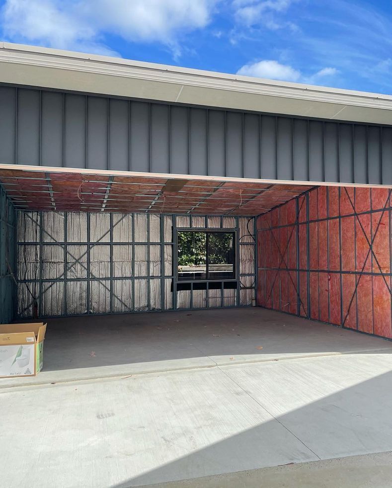 Open hangar interior, metal frame walls, insulation, window in the back, gray concrete floor, blue sky.
