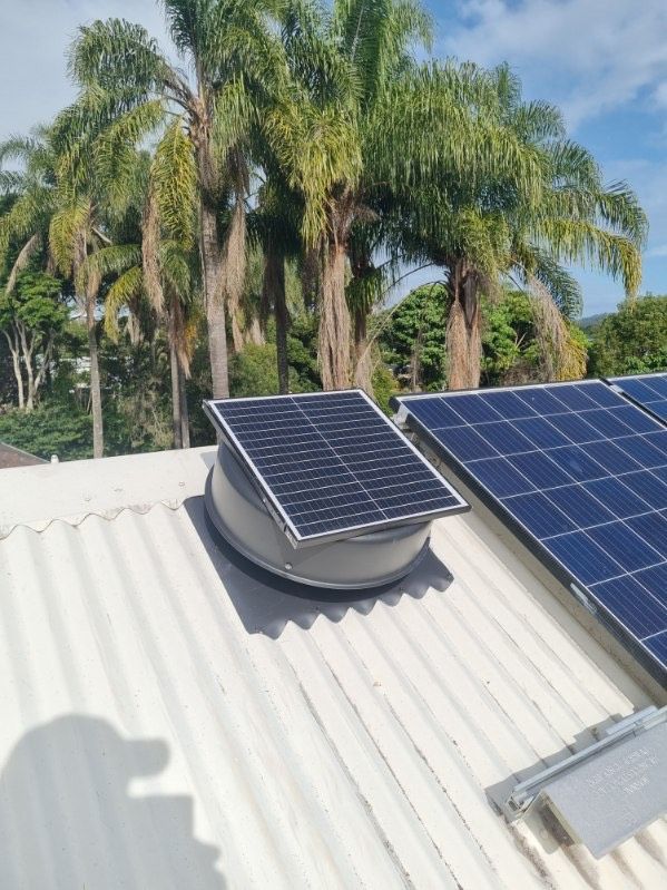 Solar-powered roof fan with solar panels on a corrugated metal roof, with palm trees in the background.