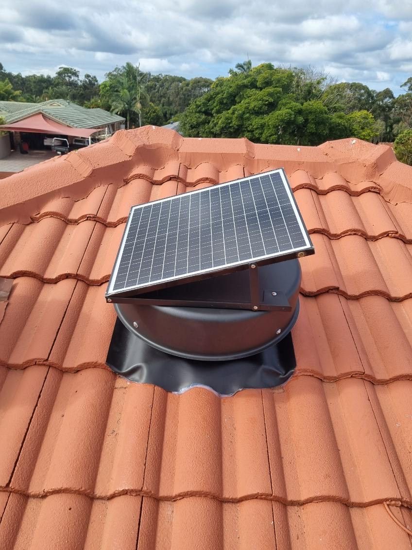 Solar-powered roof vent installed on a red-tiled roof, with a solar panel on top. Blue sky and trees in the background.