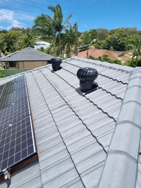 Gray tiled roof with two black turbine vents and solar panels. Trees and blue sky background.