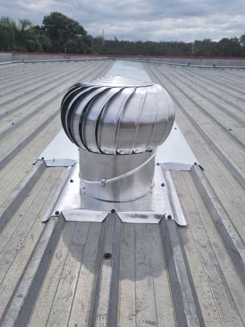 A silver, spinning turbine vent on a corrugated metal roof under a cloudy sky.