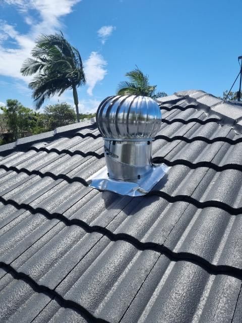 Shiny, metal roof turbine vent on a gray tiled roof under a blue sky with palm trees.