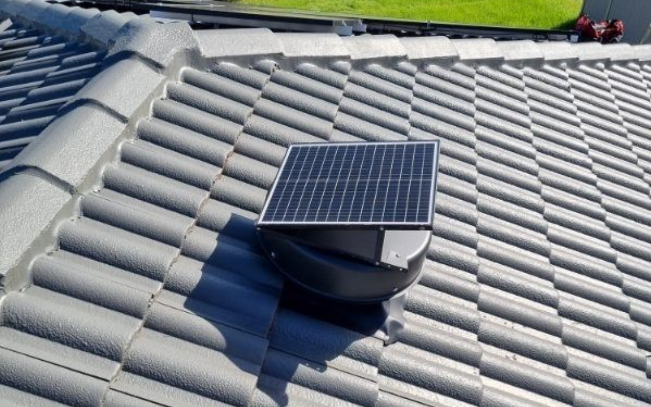 Solar-powered roof vent on a gray tiled roof. The solar panel is square and mounted above the vent.