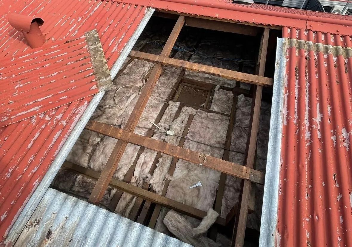View of a damaged red corrugated roof with a hole exposing insulation and wooden support beams.