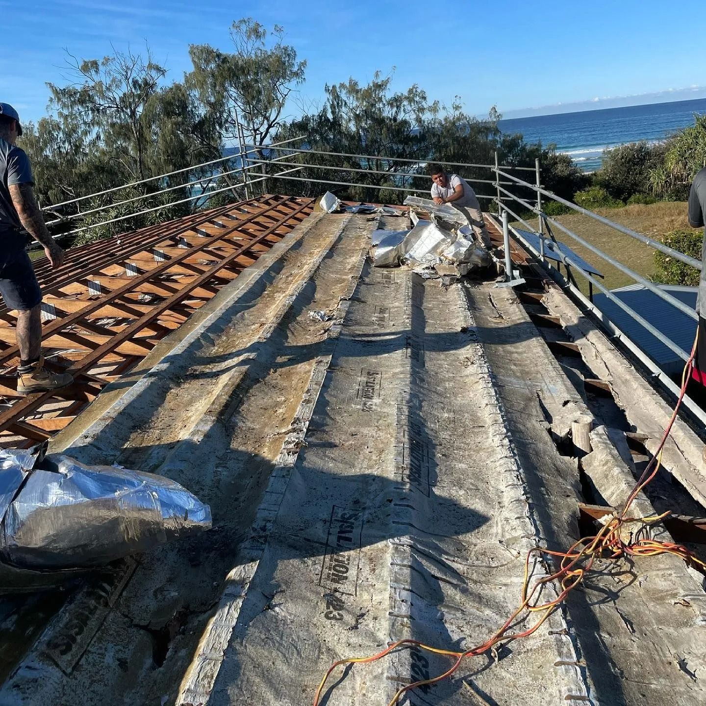 Workers on a roof removing old tiles, near the ocean. Scaffolding surrounds the edge.