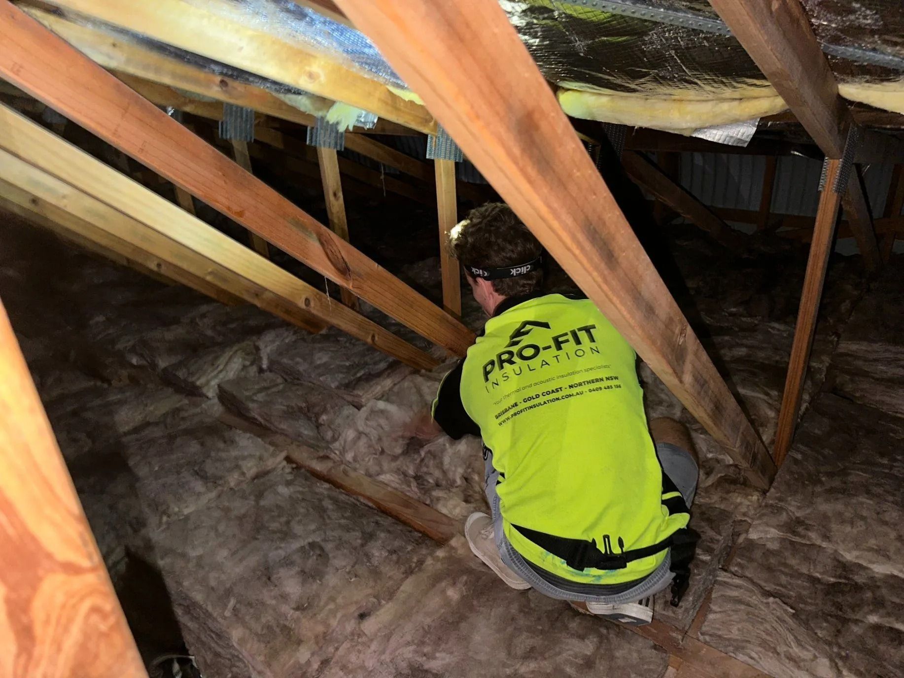 Person in yellow vest inspecting attic insulation, surrounded by wooden beams.