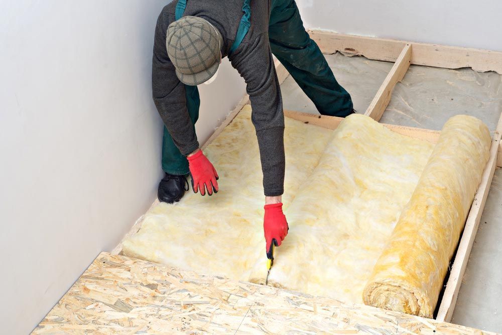 Person in red gloves cutting insulation with a utility knife, installing it between wooden floor joists.