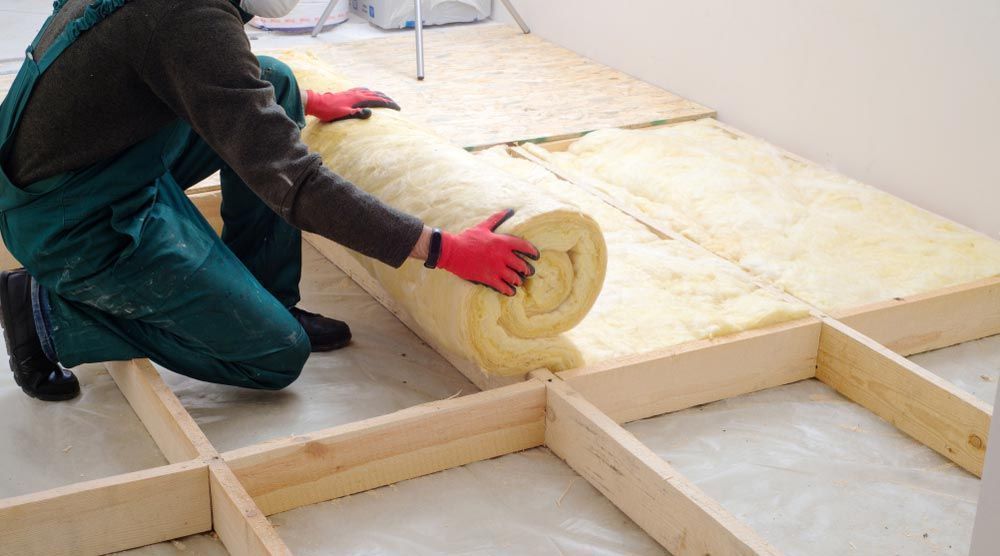 Person in green overalls installing insulation roll between wooden floor beams.