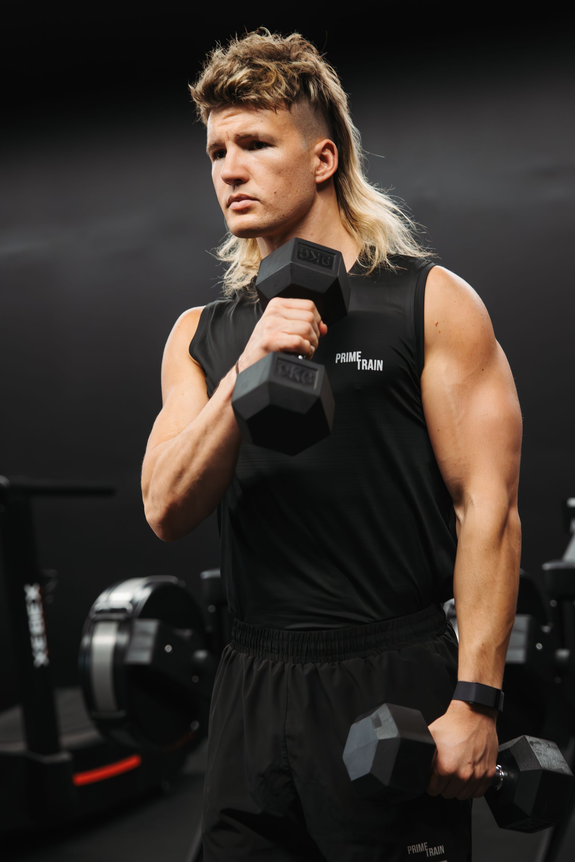 A man with a mullet is holding two dumbbells in a gym.