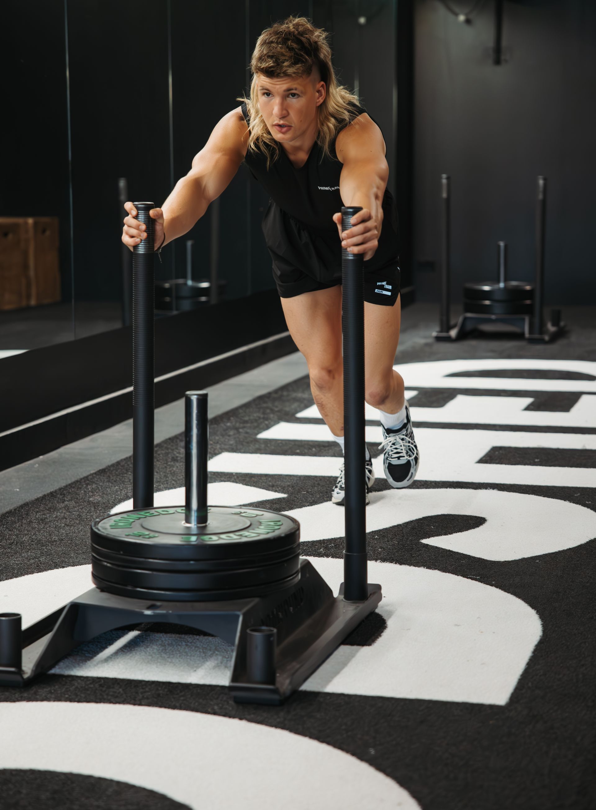 A man with a mullet is pushing a sled in a gym.