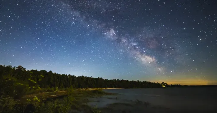 The milky way is visible over a lake with trees in the foreground.