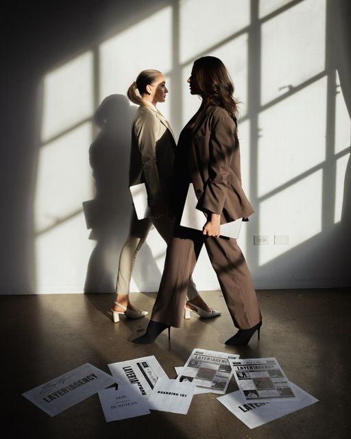 Two women in suits face each other, backlit by sunlight, papers on the floor.
