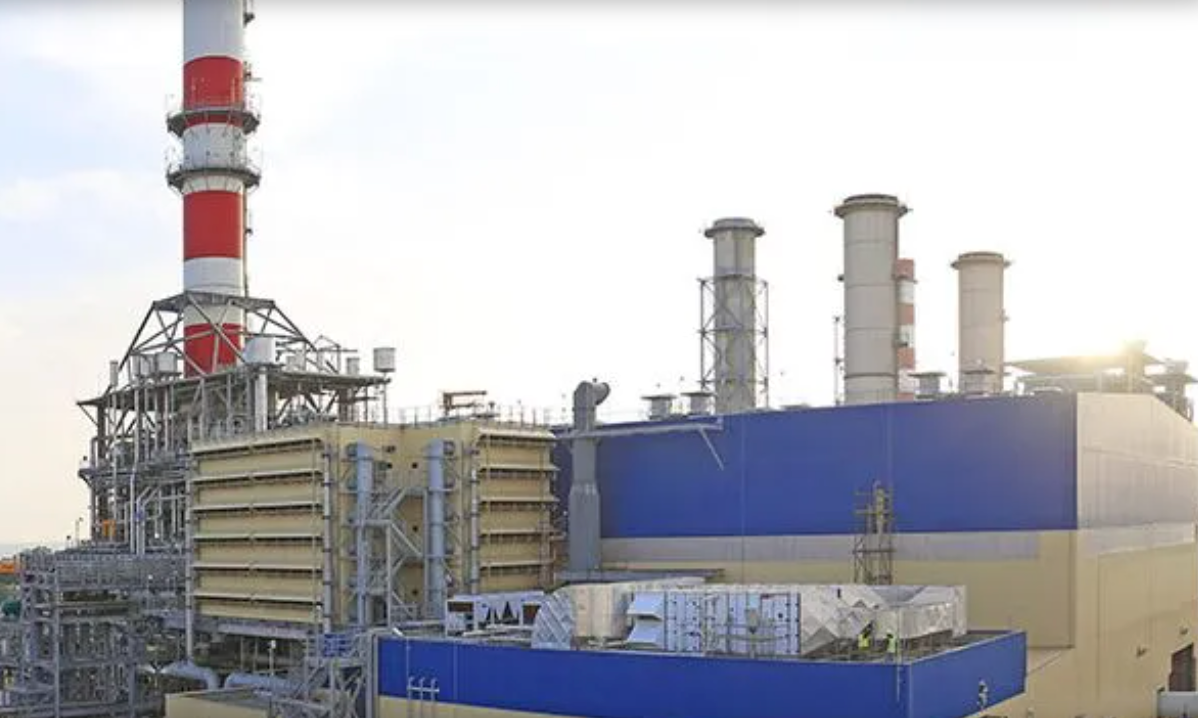 A panoramic view of an industrial plant with red and white smokestacks against a blue sky.