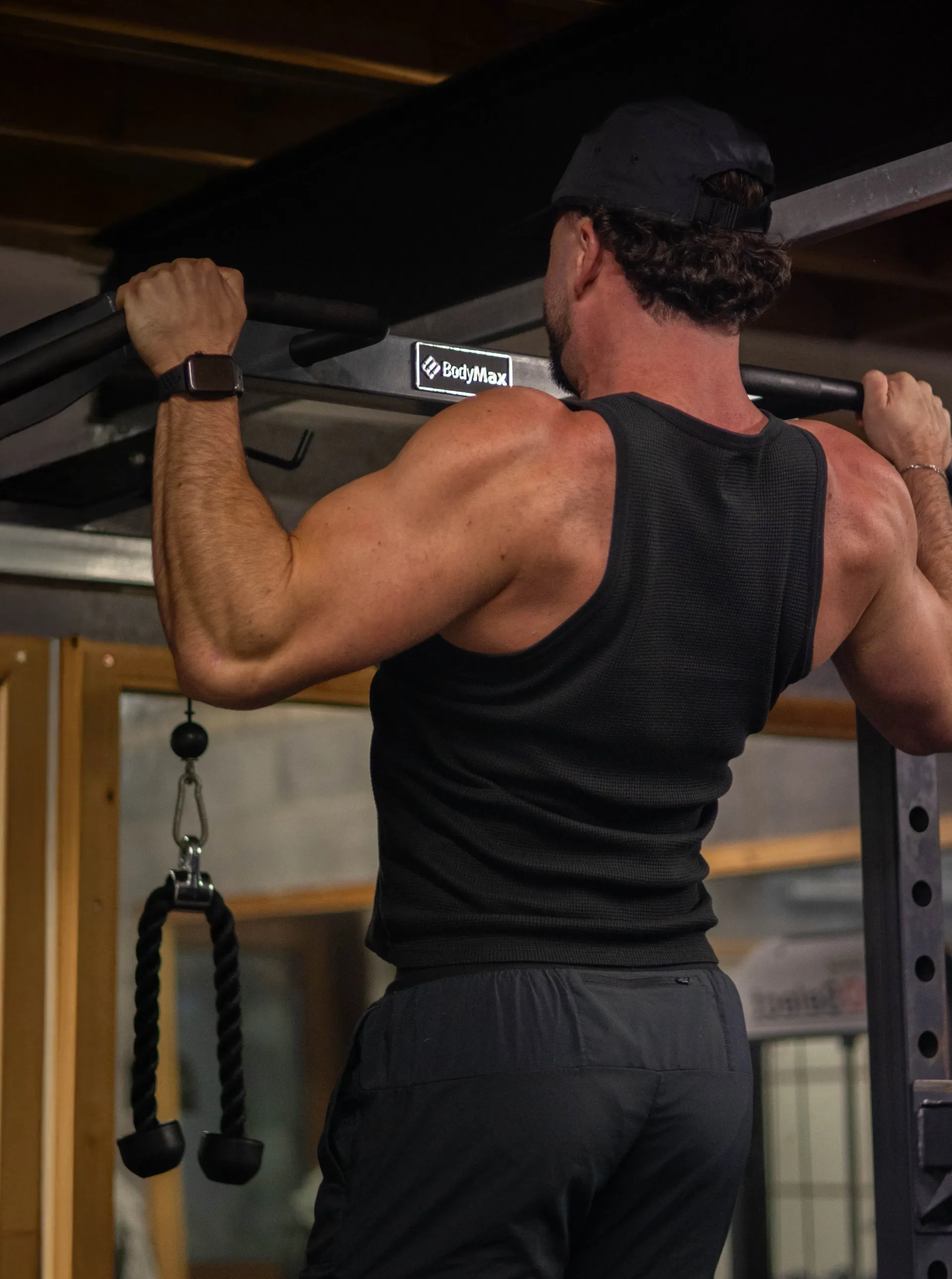 Man doing a pull-up, wearing a black tank top and hat in a gym.