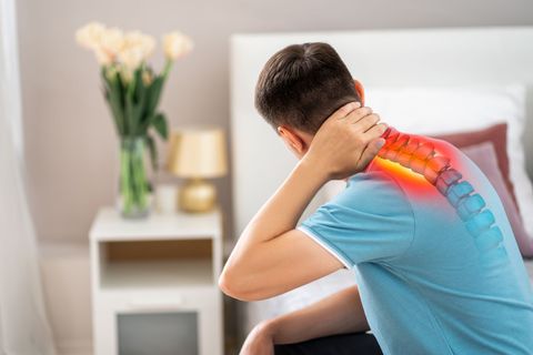 Man seated, hand on neck, spine highlighted with red, indicating pain, in a bedroom.