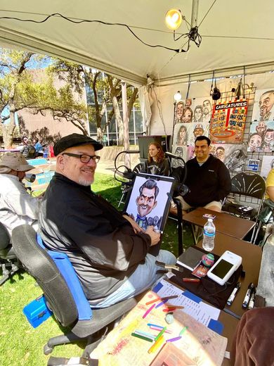 People seated under a white tent at an outdoor art booth, with colorful drawings displayed on the table.