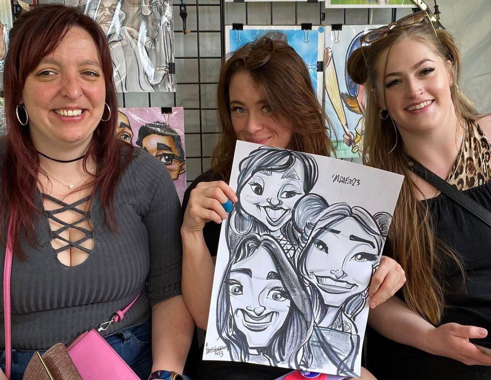 Three smiling people at a booth, one holding a black-and-white caricature drawing.