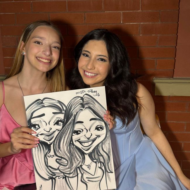 Two smiling friends holding a caricature drawing in front of a brick wall