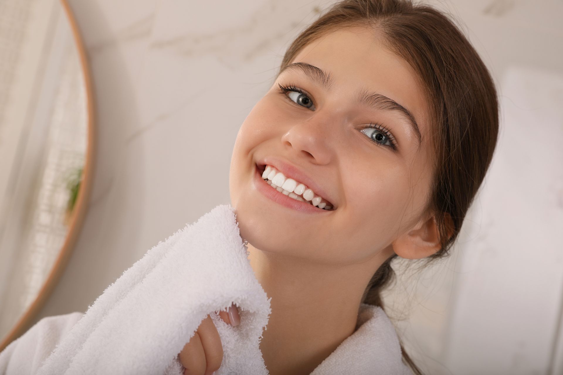 A smiling person in a white robe holds a white towel against their neck in a bright, modern bathroom.