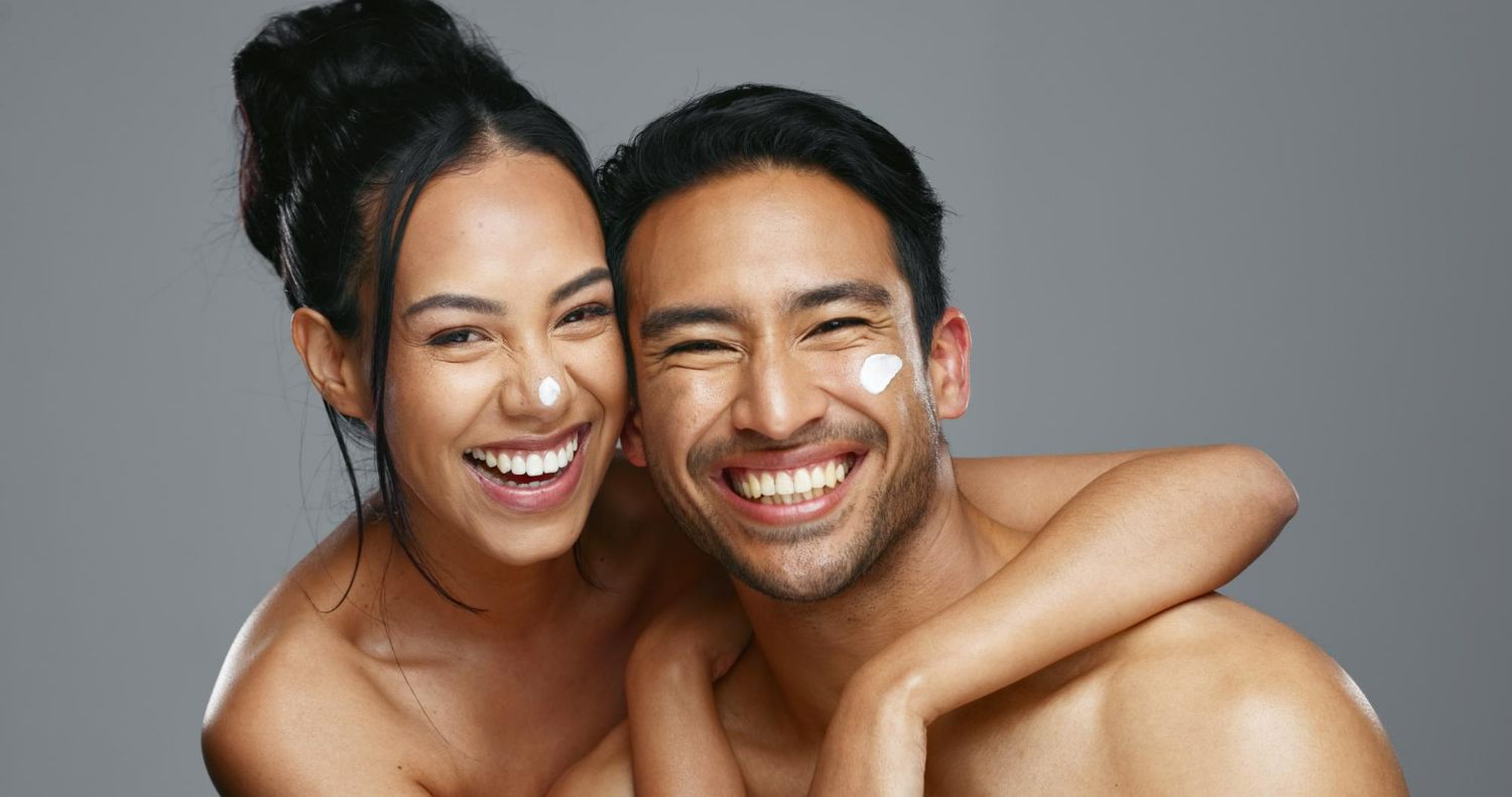 Smiling woman embraces a smiling man with a heart of cream on his cheek, both bare-chested against a gray background.