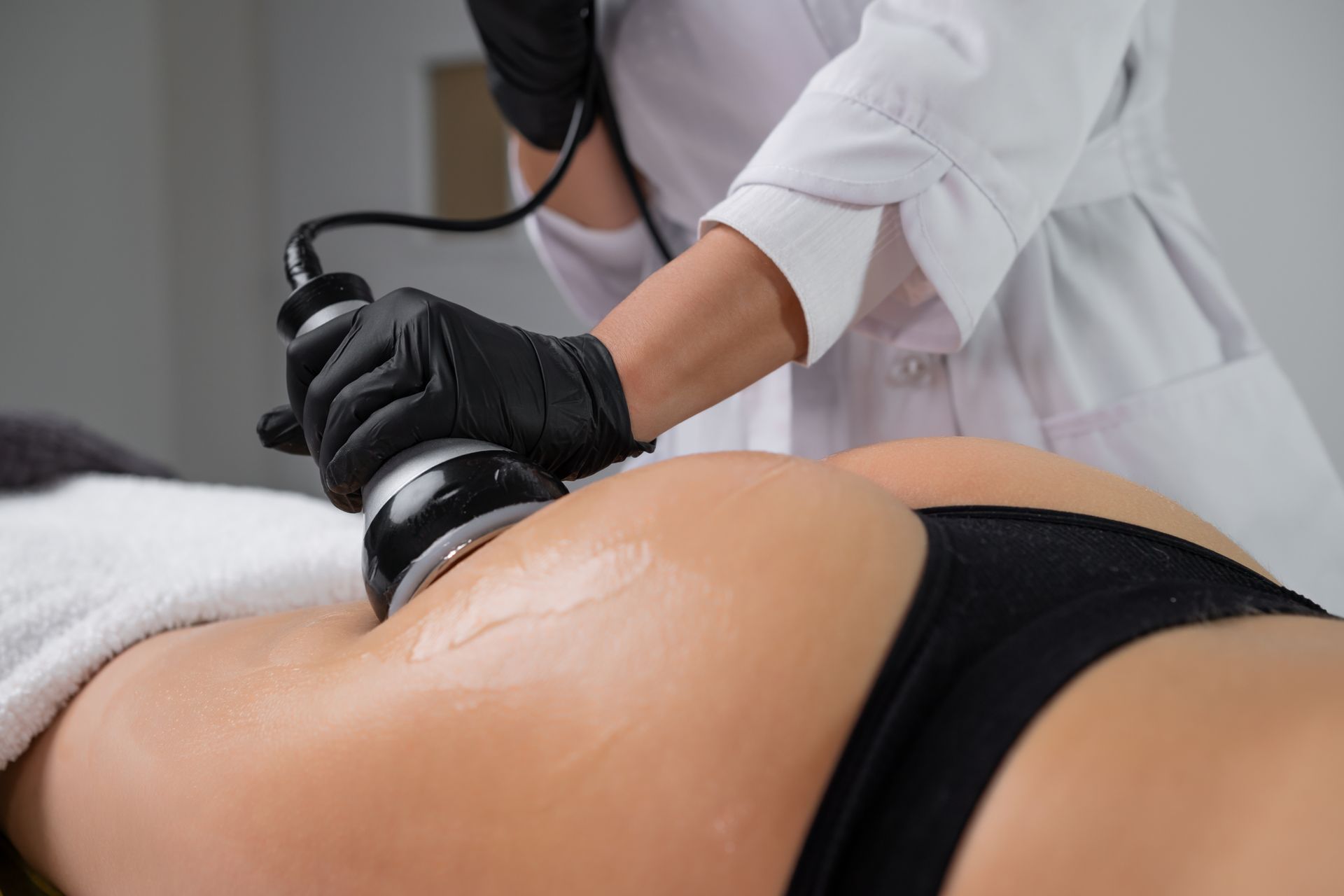A practitioner in a white coat uses a cosmetic handheld device on a client's gluteal area in a spa setting.