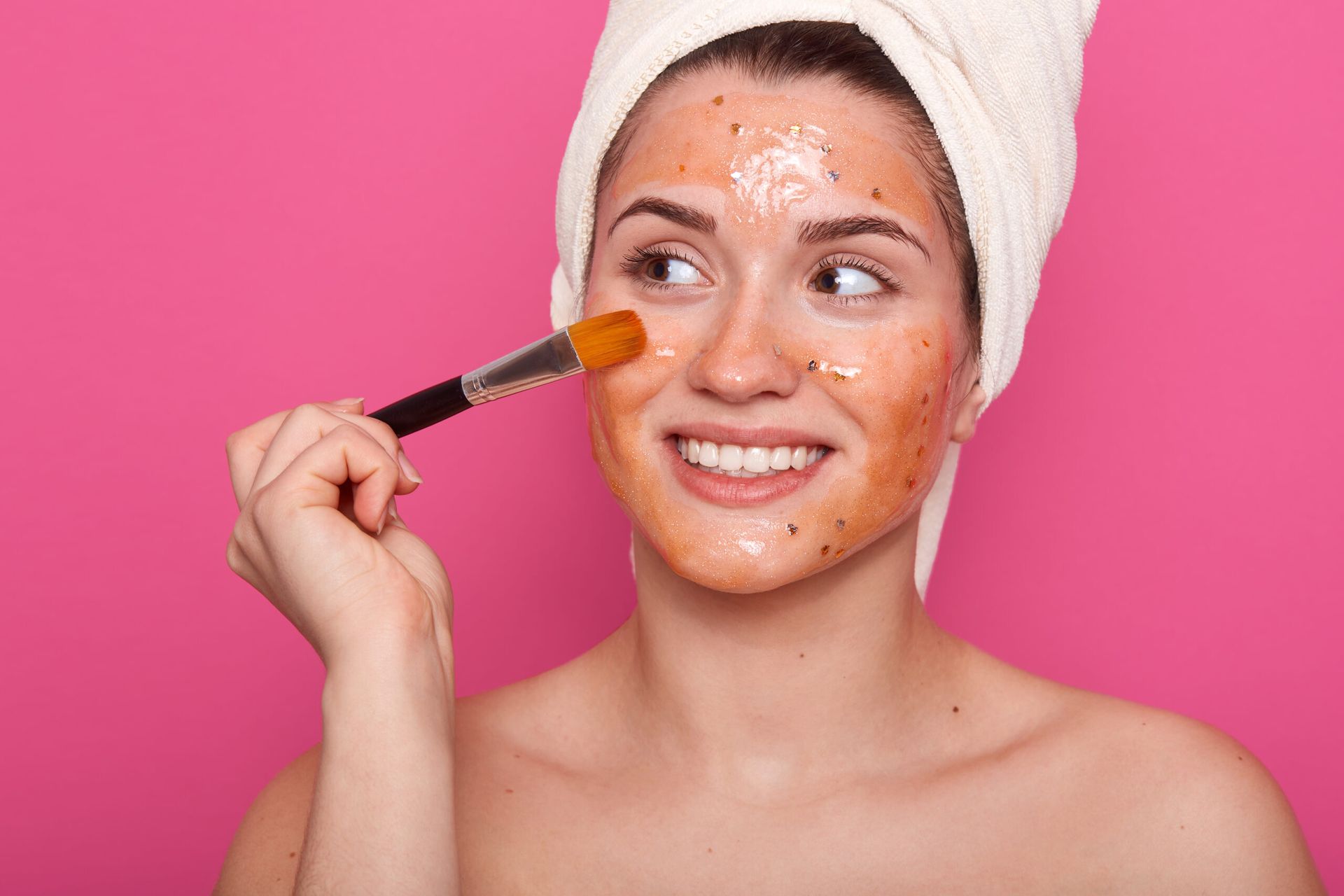 A woman with a towel wrapped around her head is applying a mask to her face.