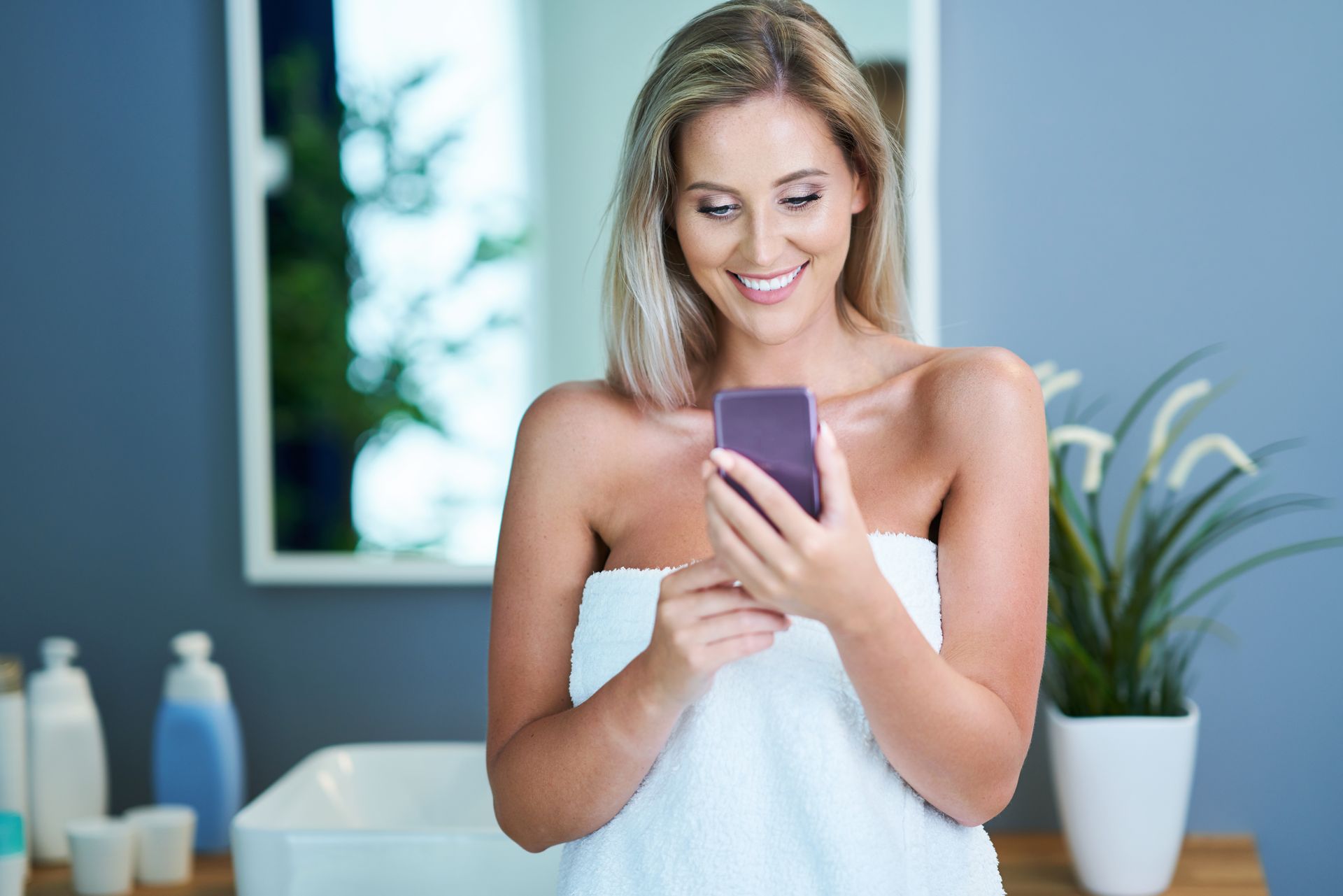 A woman wrapped in a towel is using a cell phone in a bathroom.