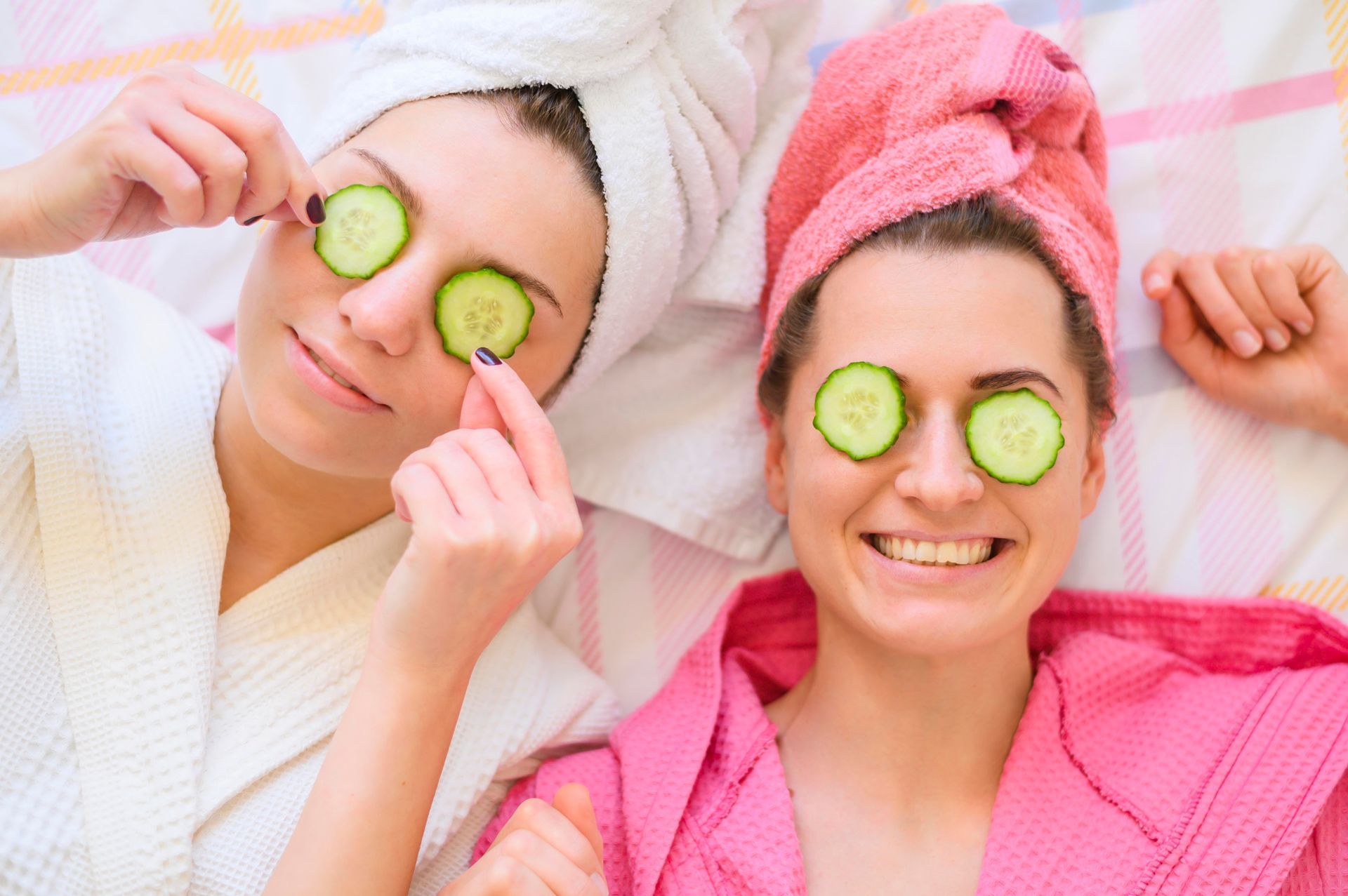 Two women are laying on a bed with cucumber slices on their eyes.