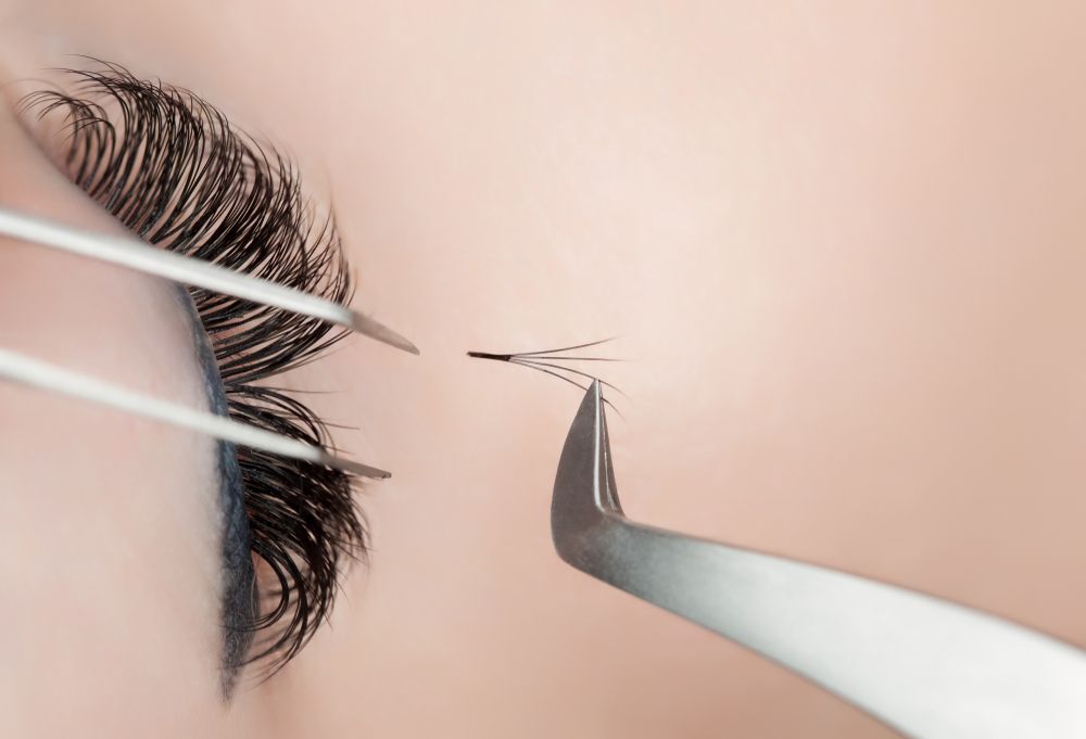 Close-up of eye with lash extensions being applied with tweezers. Skin is light pink.
