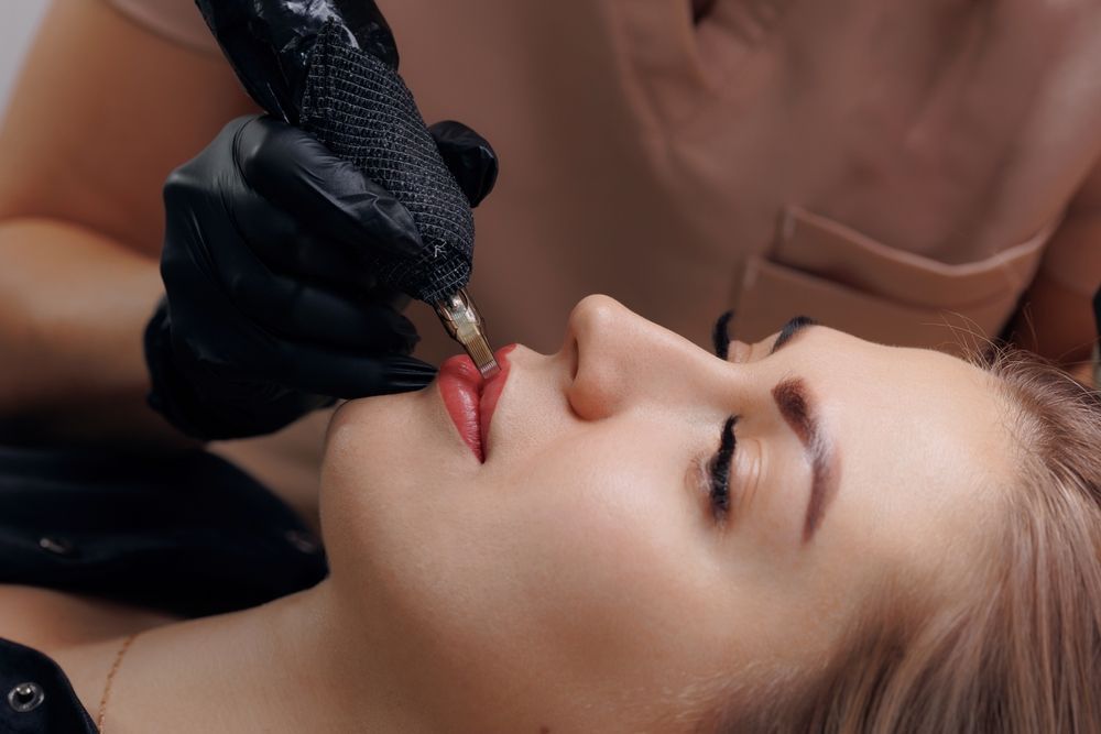 Person receiving lip tattooing procedure, close-up with artist's hand, black gloves, and tattoo machine.
