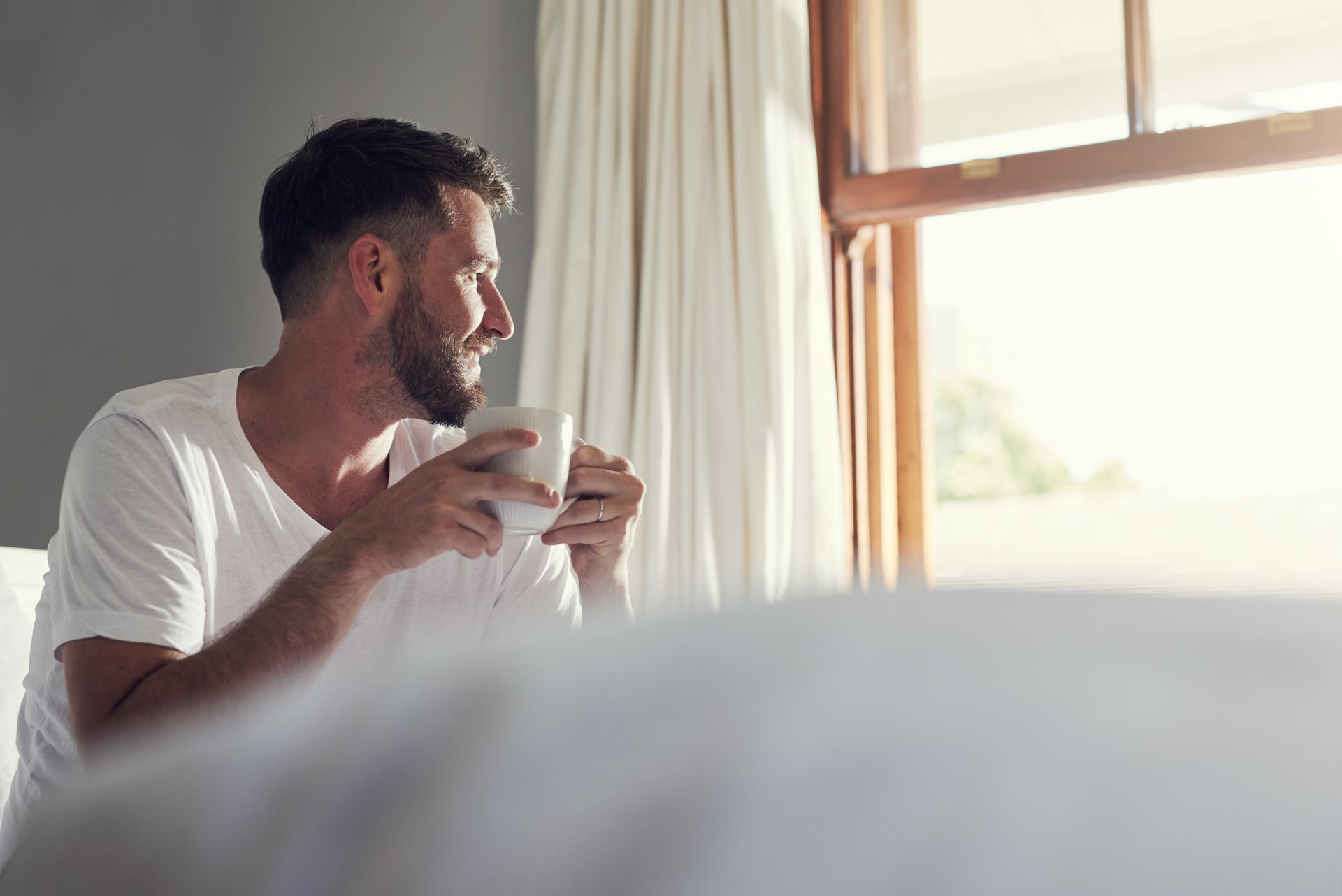 man waking up from bed holding a cup in his hand, with the morning sunlight streaming through a wind