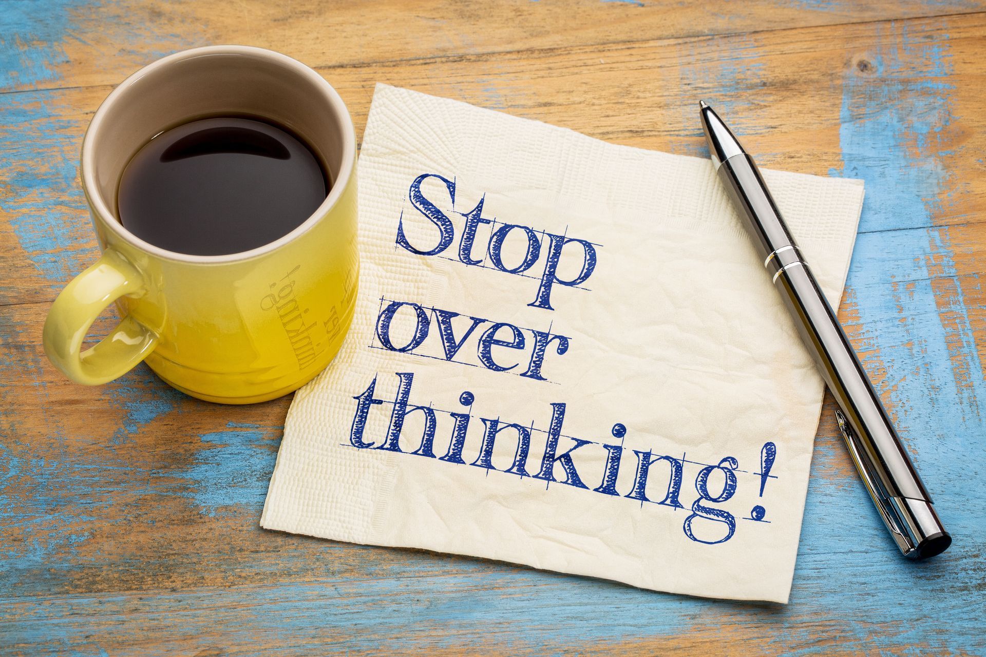 Coffee cup beside note reading “Stop over thinking!” on a wooden table with a pen