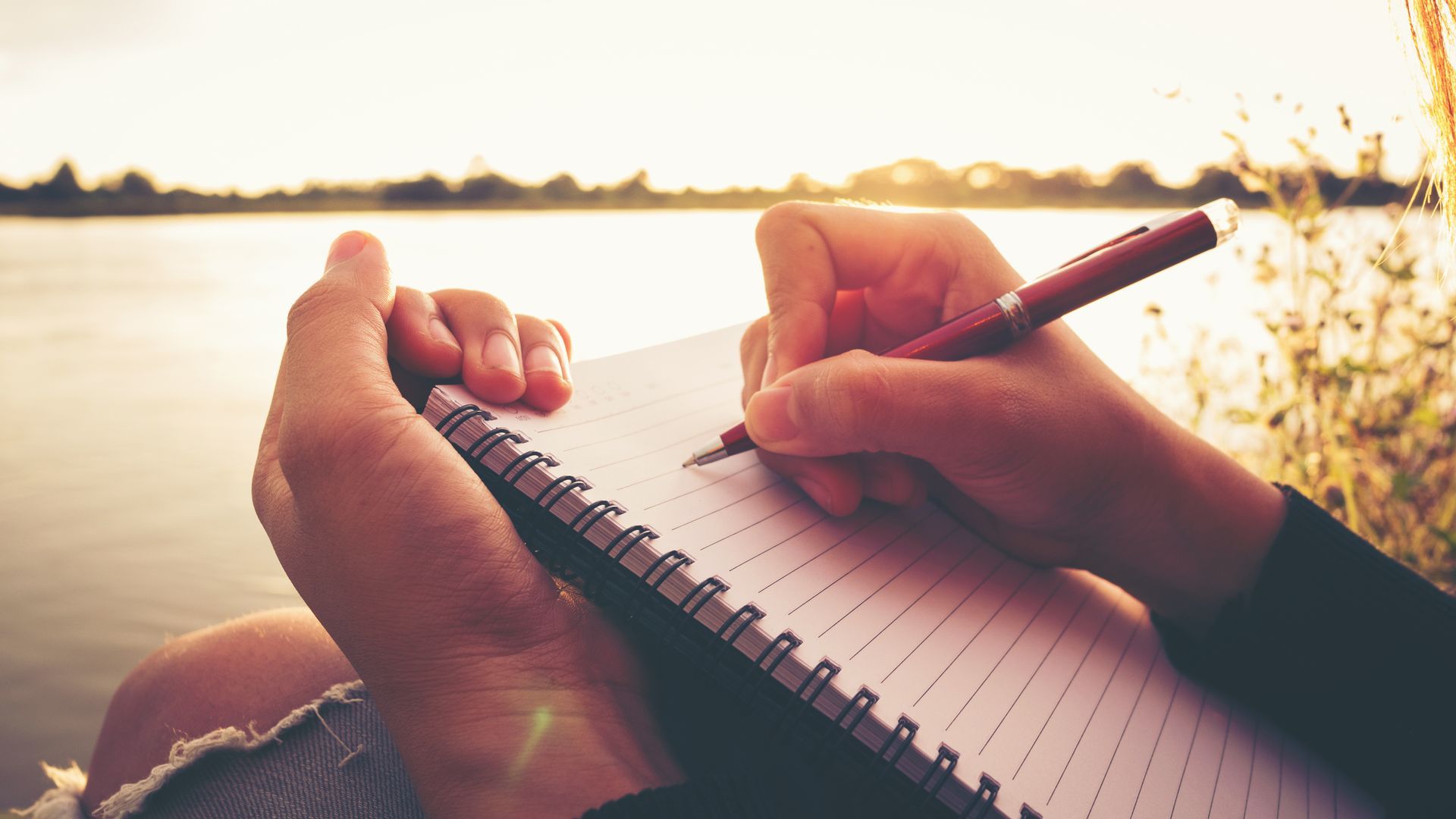 Hands writing in a spiral notebook by a bright waterfront at sunset.