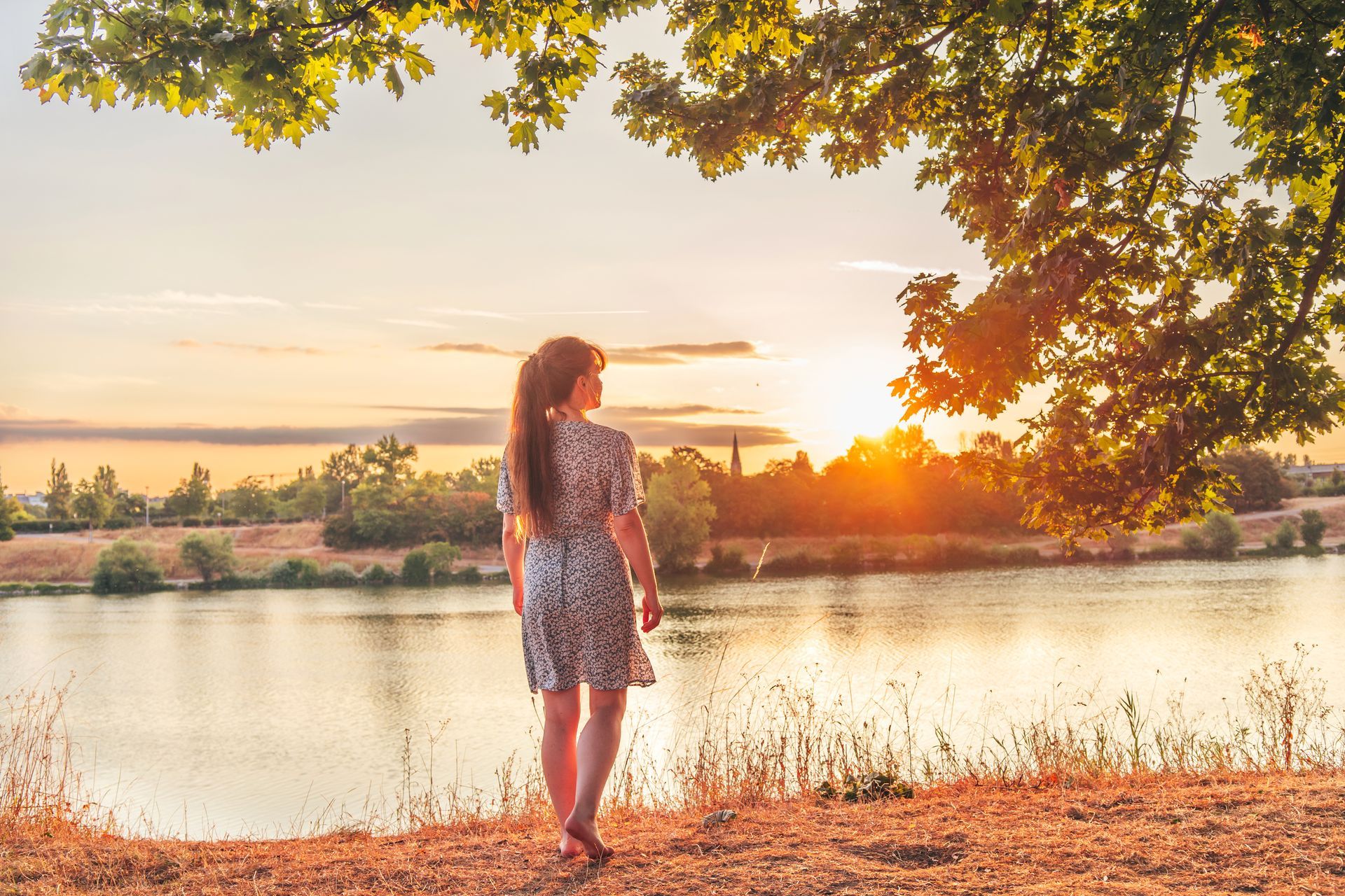 A person with long hair stands by a river at sunset, framed by golden sunlight and overhanging tree branches.