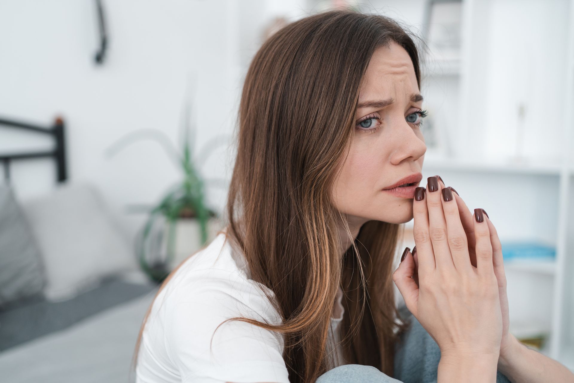 A person with a worried expression sits with their hands clasped together near their mouth in a bedroom setting.