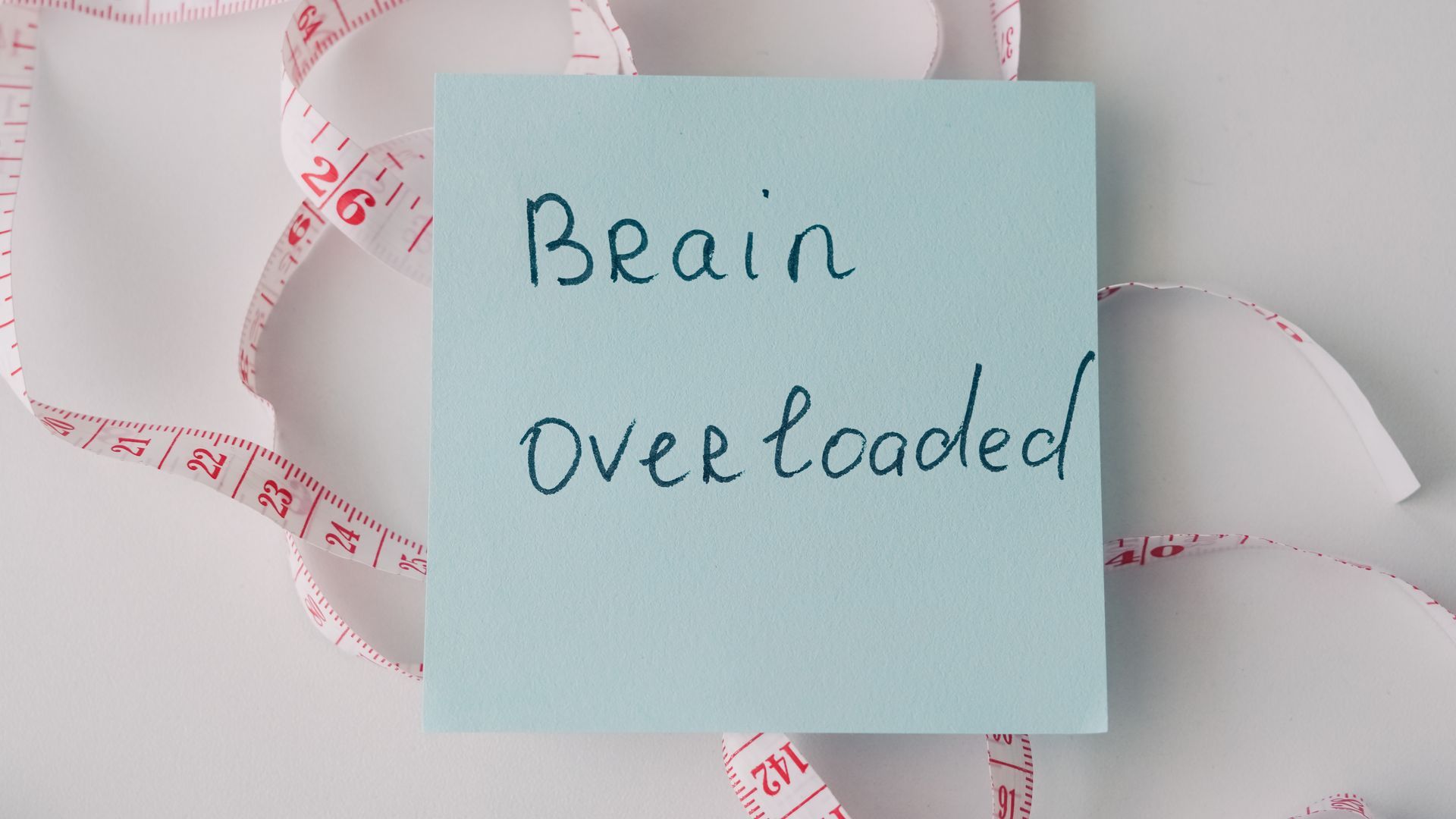 Blue sticky note reading “Brain overloaded” on a white background with a measuring tape