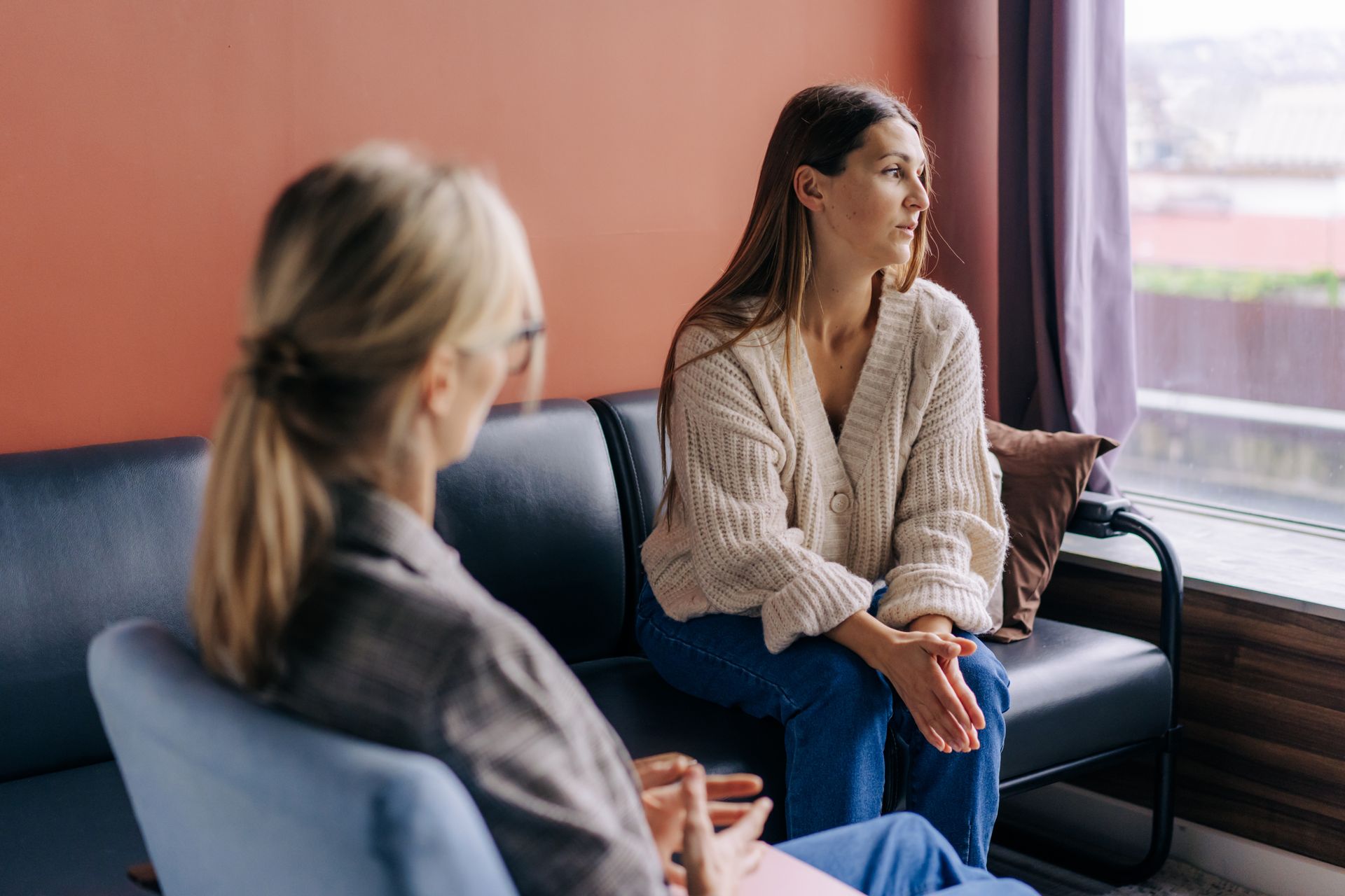 Two women sit on a couch by a window, talking in a calm, supportive setting.