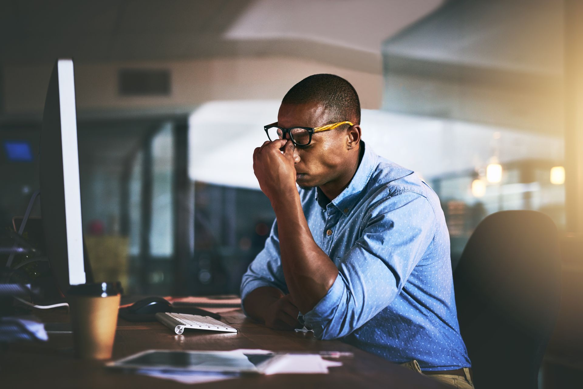 A professional wearing glasses sits at a desk, rubbing their eyes in a dimly lit office while working at a computer.