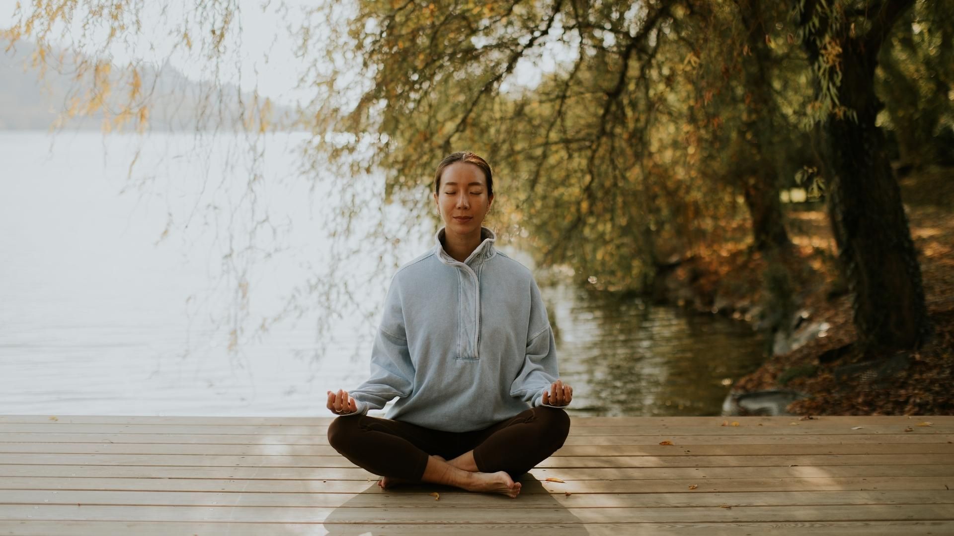 Person meditating cross-legged on a wooden dock by a calm lake, with trees and autumn foliage in the background.