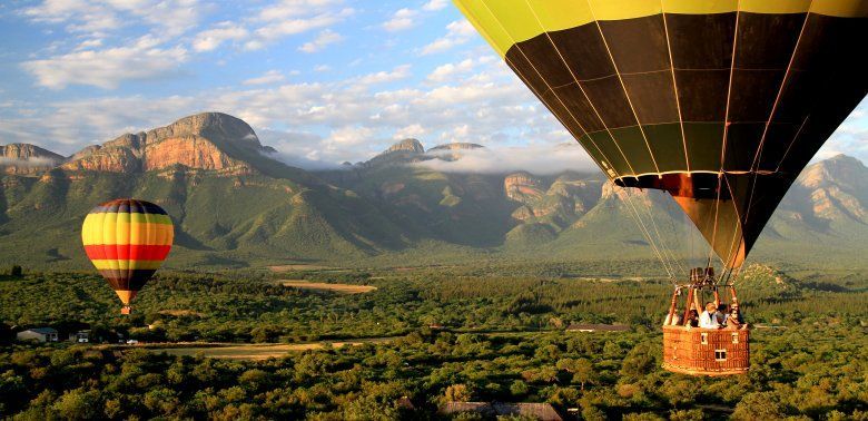 Two hot air balloons are flying over a lush green valley with mountains in the background.