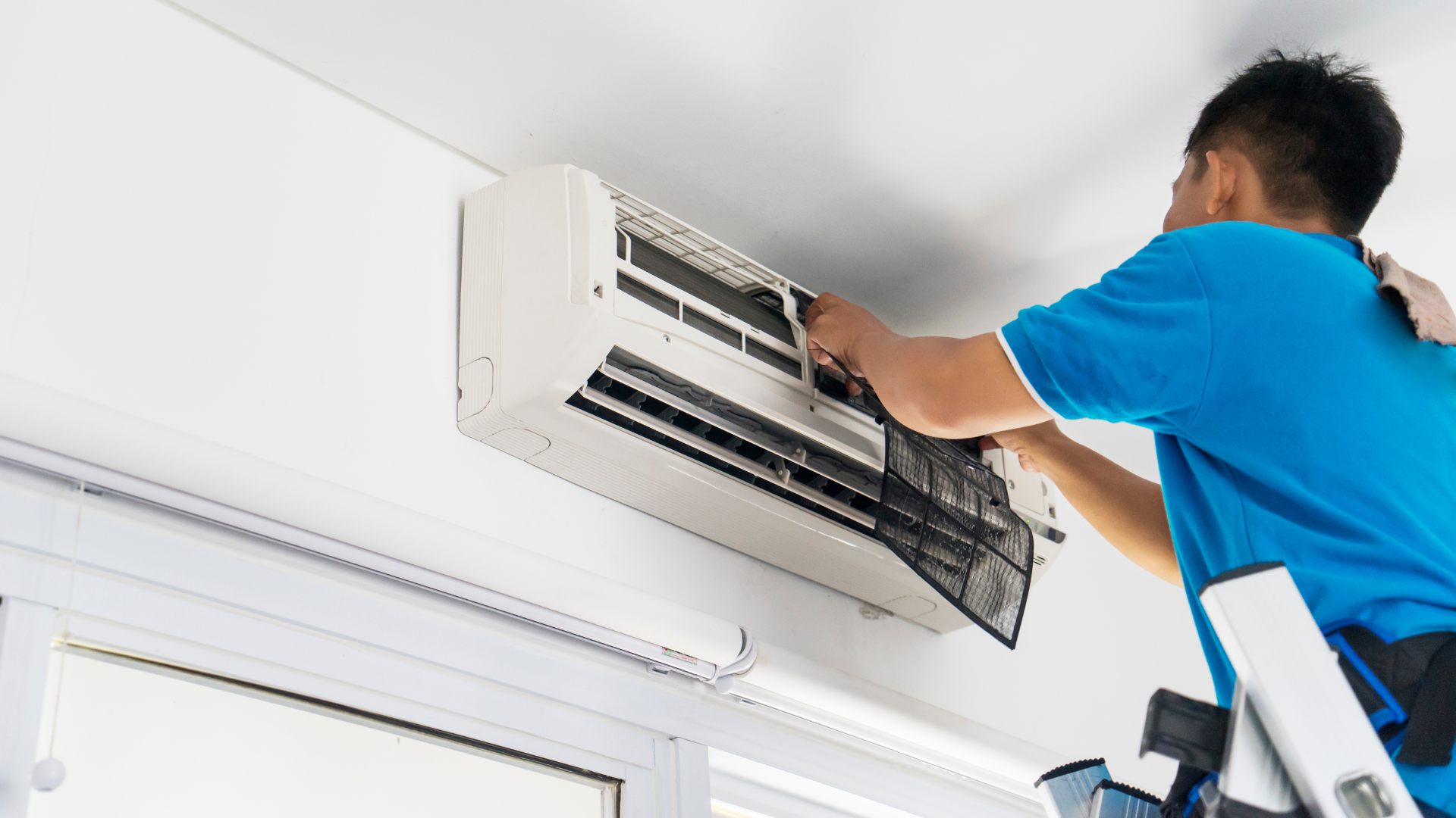 A man is standing on a ladder fixing an air conditioner.