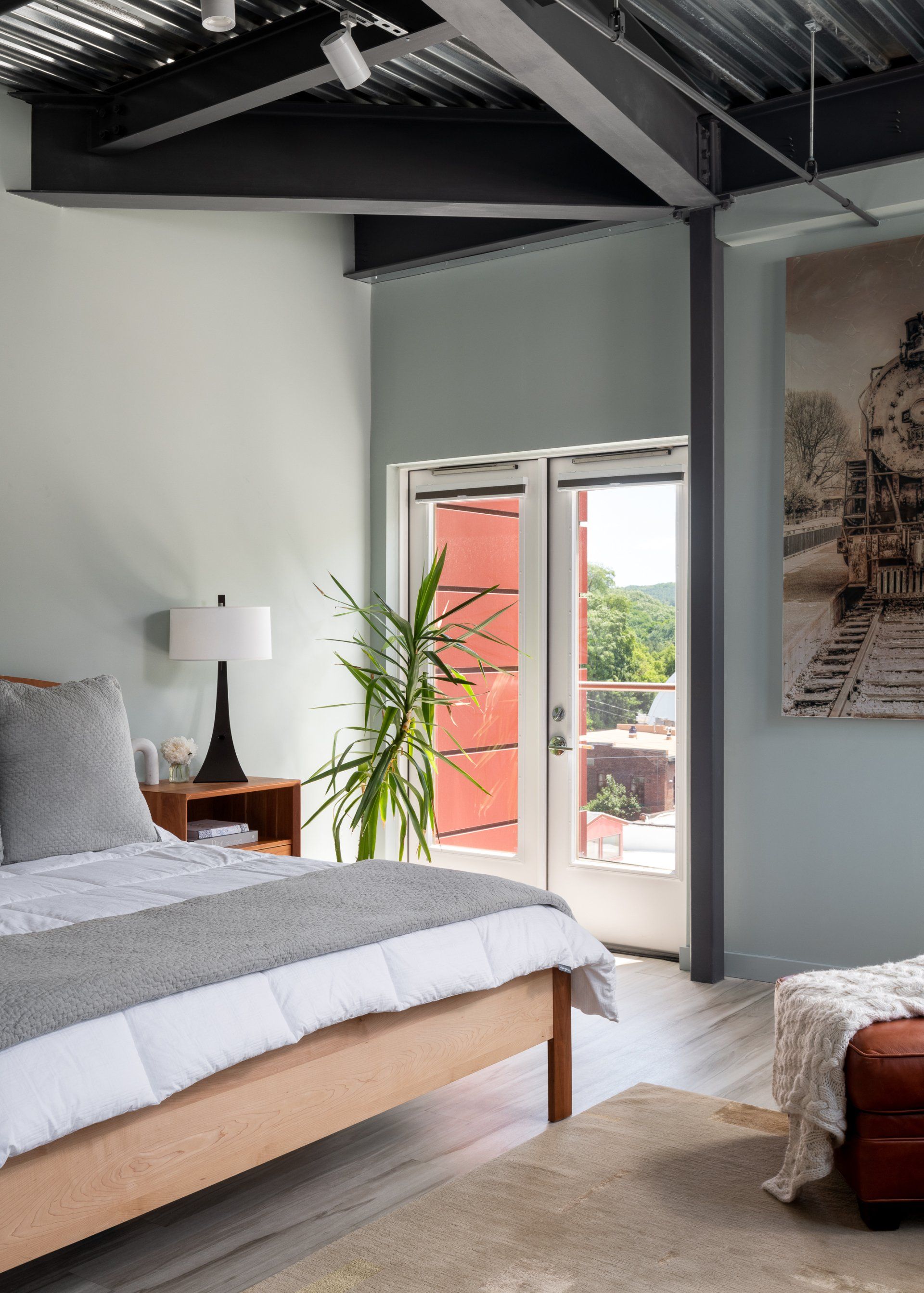 A bedroom with a bed , lamp , ottoman and a painting on the wall.