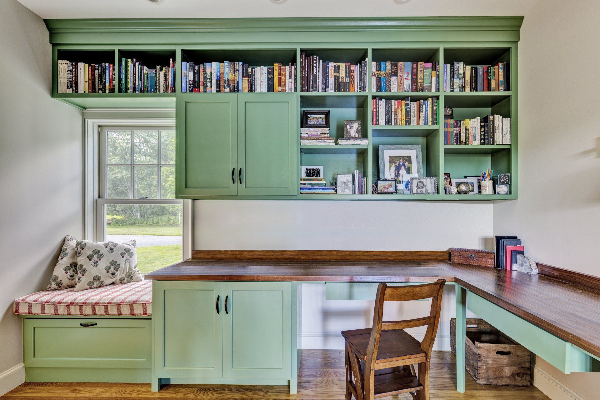 A room with green cabinets , a desk , and a window seat.