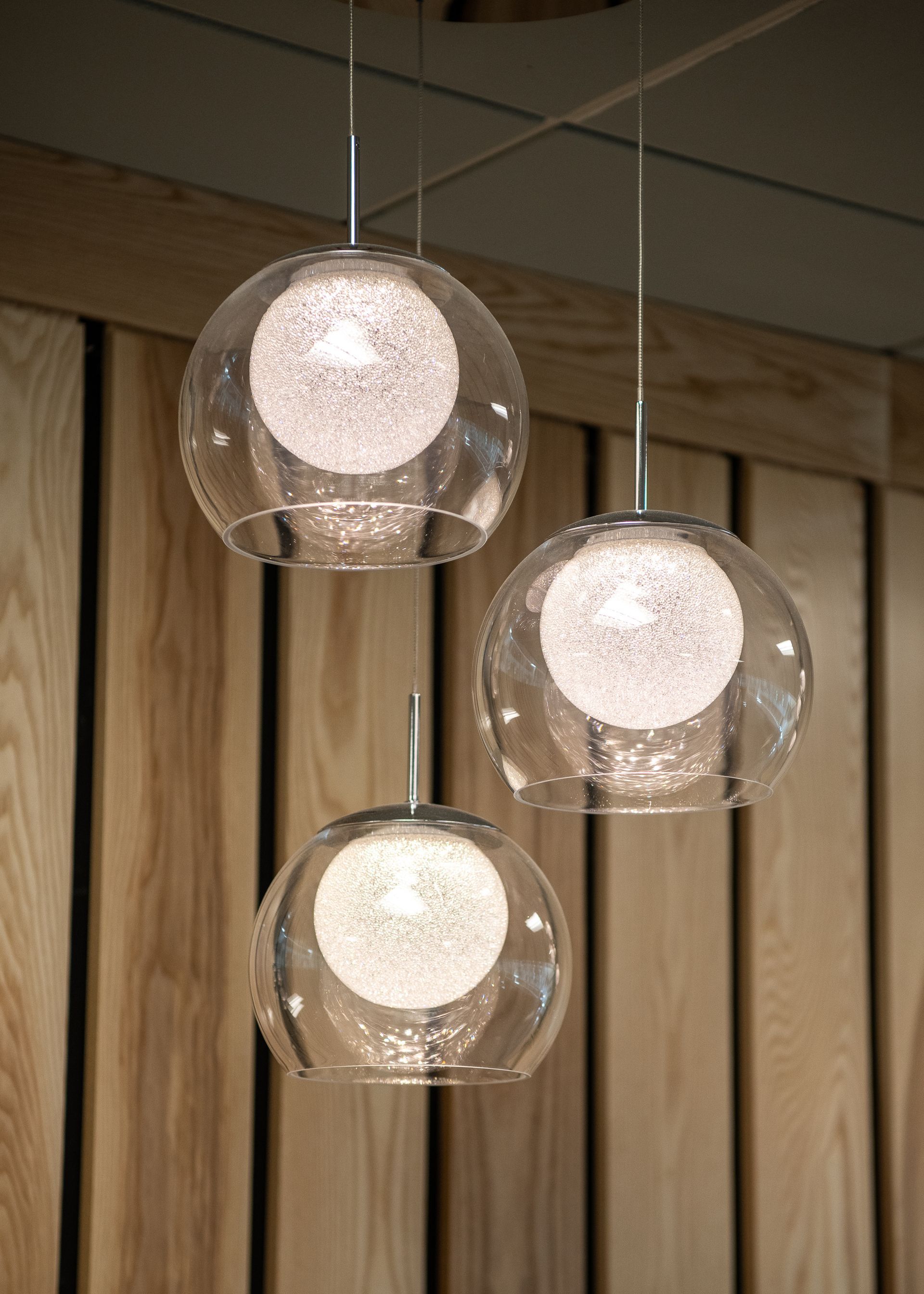 Three clear glass pendant lights are hanging from the ceiling in front of a wooden wall.