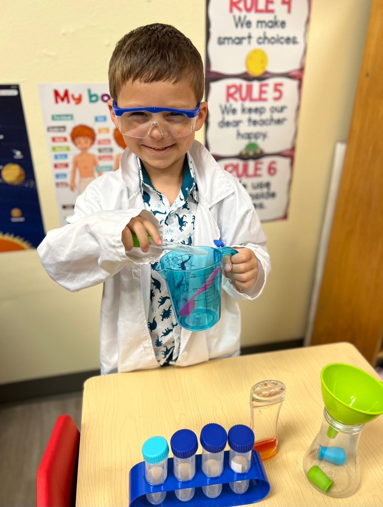 a young boy in a lab coat and goggles is pouring liquid into a measuring cup .