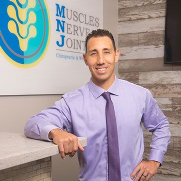 Man in purple shirt and tie smiling in a clinic lobby beside a “Muscles Nerves Joints” sign